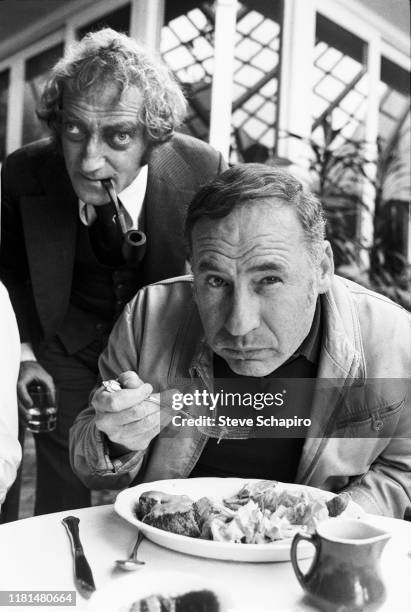 Pipe in his mouth, English actor and comedian Marty Feldman stands behind film director, actor, and comedian Mel Brooks, the latter of whom eats a...