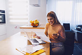 Cute caucasian smiling blonde woman in sweater holding bills in one hand and in other smart phone. On table are laptop and bills. Apartment interior.