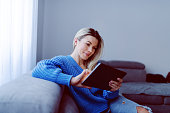 Beautiful caucasian smiling blonde in blue sweater and jeans sitting on sofa in living room and using tablet. Apartment interior.