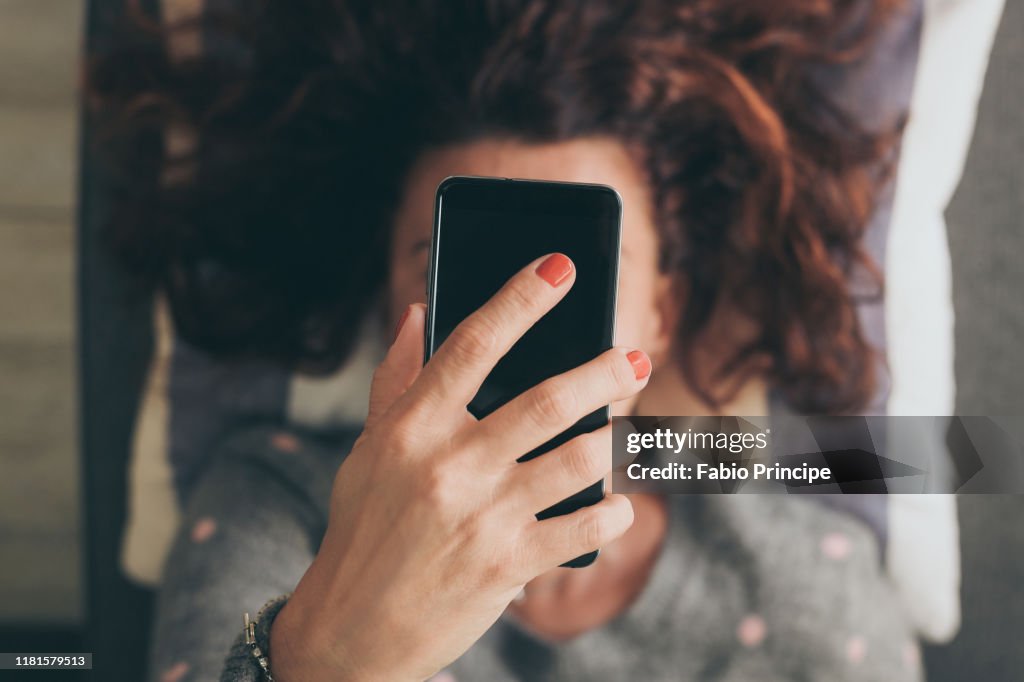 Frau hält Handy über Gesicht, während sie auf dem Sofa liegt