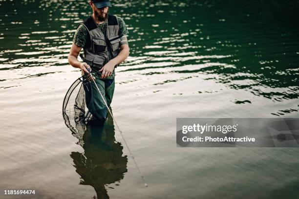 Man Wearing Waders Standing In A River Holding Large Fish Net With