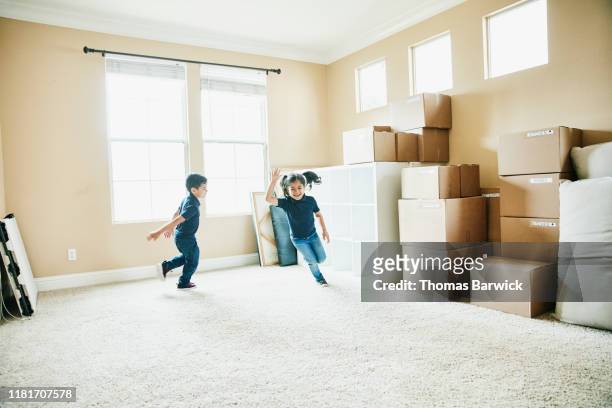 Young brother and sister running in living room filled with boxes on moving day