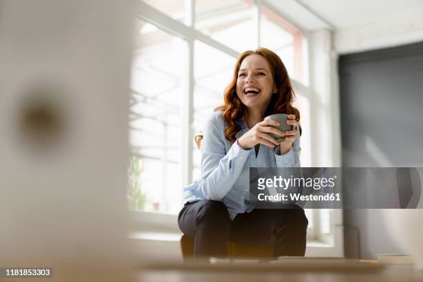 portrait of laughing redheaded woman with tea bowl sitting on backrest in a loft - blue blouse - fotografias e filmes do acervo