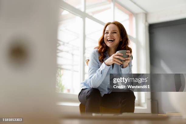 portrait of laughing redheaded woman with tea bowl sitting on backrest in a loft - laughing fun bildbanksfoton och bilder