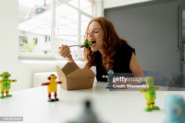 portrait of redheaded businesswoman in a loft with miniature robots on desk eating healthy takeaway food - virtual lunch stock pictures, royalty-free photos & images