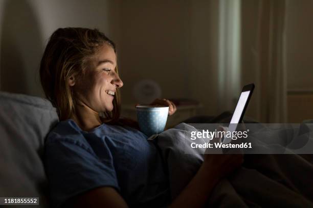 young woman lying in bed at home with cup of tea at night using tablet - e reader stock-fotos und bilder