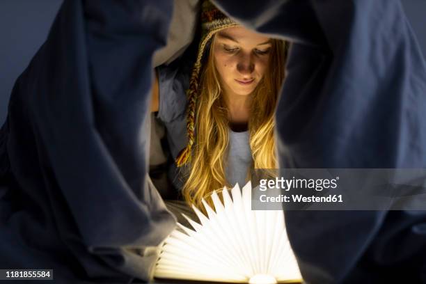 young woman reading illuminated book in bed at home - rechtschreibung stock-fotos und bilder