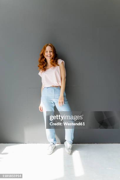 portrait of laughing redheaded woman standing in front of grey wall - ganzkörperansicht stock-fotos und bilder