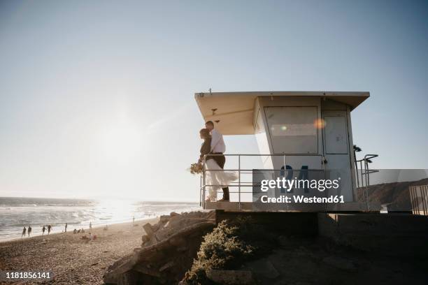 bride and groom at lifeguard hut on the beach at sunset - sunset beach bouquet wedding couple stock pictures, royalty-free photos & images