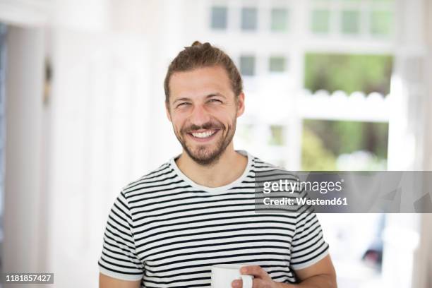 portrait of smiling young man holding cup of coffee at home - solo un uomo giovane foto e immagini stock