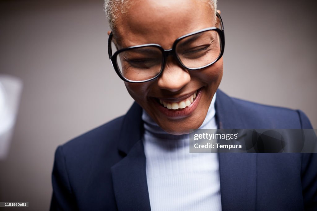 Afro Caribbean woman, dressed in a suit and wearing glasses, smiling and shy.
