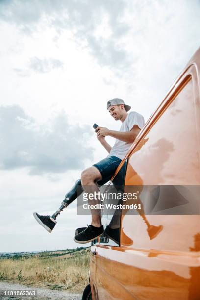 young man with leg prosthesis sitting on roof of camper van using cell phone - equipo protésico fotografías e imágenes de stock