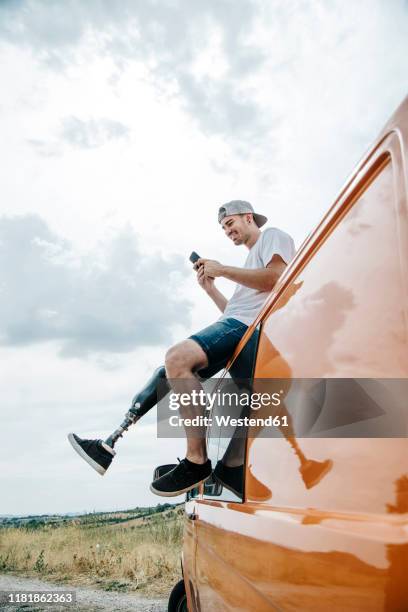 young man with leg prosthesis sitting on roof of camper van using cell phone - medical technology stock pictures, royalty-free photos & images