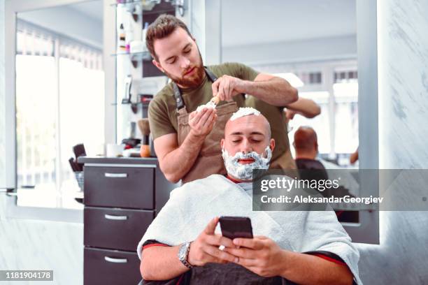 barber puts shaving cream on relaxed customer while he's using smartphone - beard trimmer stock pictures, royalty-free photos & images