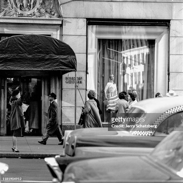 Outside of the Bergdorf Goodman clothing store, a woman in a black fur hat and fur-trimmed coat hails a taxi as pedestrians walk past and window...