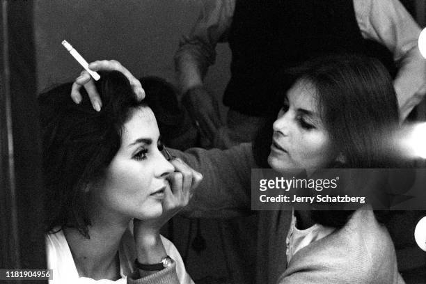 American actress Millie Perkins sits as model Anne St Marie , a cigarette in one hand, applies Perkin's eye makeup, New York, New York, 1963.