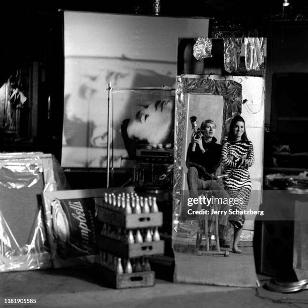 In his studio, the Factory, American Pop artist Andy Warhol sits on a stationary bicycle with a movie camera in his hands while beside him stands an...