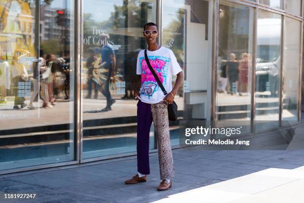 Model Kalib Besher wears reflective sunglasses, a white graffiti boom box t-shirt, duo purple and print pants, and brown leather loafer shoes after...