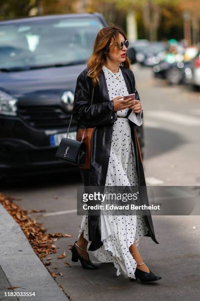 Guest wears sunglasses, a black leather long jacket, a white pleated dress with printed polka dots, a Saint Laurent YSL bag, black leather pointy...