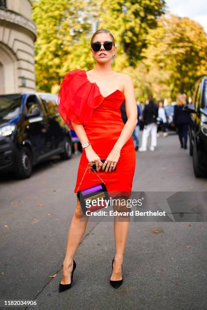 Guest wears sunglasses, earrings, a red ruffled off-shoulder dress, a purple yellow and red bag, black pointy shoes, outside Elie Saab, during Paris...