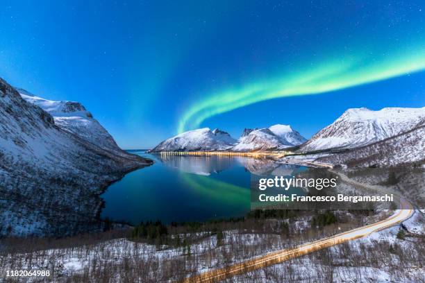 the aurora borealis lights up in the sky and is reflected in the fjord with a road in the foreground. northern norway, europe. - norway landscape stock-fotos und bilder