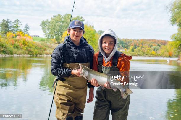 brothers holding a freshly caught rainbow trout or steelhead on a fall day. - kids at river stock pictures, royalty-free photos & images