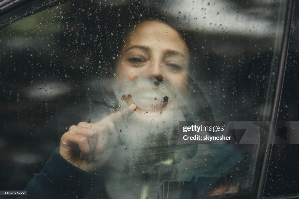 Mujer feliz dibujando la cara sonriente en la ventana del coche durante el día lluvioso.