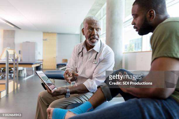 männlicher orthopädischer arzt zeigt übungen für patienten auf digitaler tablette - black men feet stock-fotos und bilder