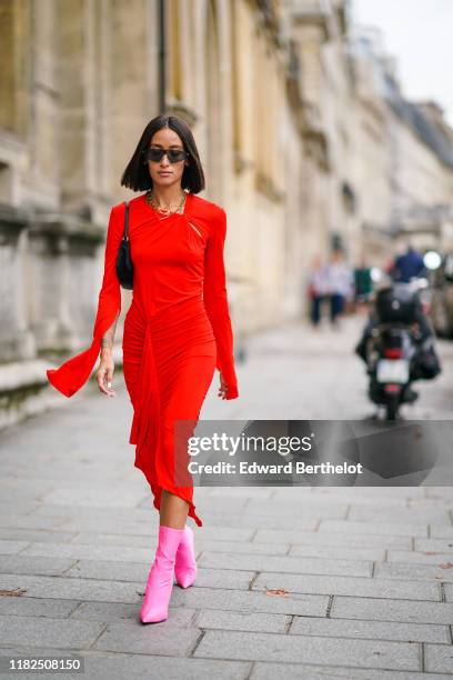 Alexandra Guerain wears sunglasses, necklaces, a black bag, a long sleeves gathered asymmetric red dress, neon-pink mid-calf sock-boots, outside...