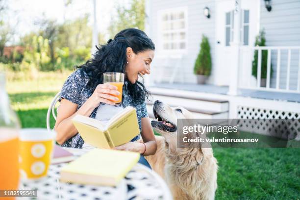 smiling mid adult woman sitting with her golden retriever on front yard and reading a book - woman playing squash stock pictures, royalty-free photos & images