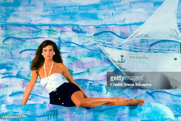 Portrait of American model and actress Carol Alt, in a two-piece swimsuit with a wrap skirt, as she poses against a blue background , Los Angeles,...