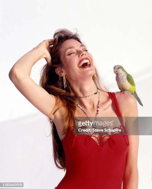 Portrait of American model and actress Carol Alt, in a red outfit, laughs as she poses with a parakeet perched on her shoulder during a photo shoot...