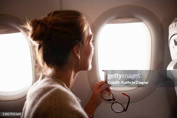 woman looking through airplane window, holding reading glasses - aeroplano foto e immagini stock