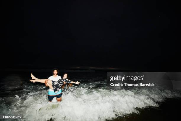 smiling man carrying woman through surf on summer evening - muscle men at beach stock-fotos und bilder