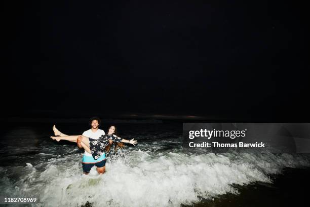 smiling man carrying woman through surf on summer evening - tough love stockfoto's en -beelden