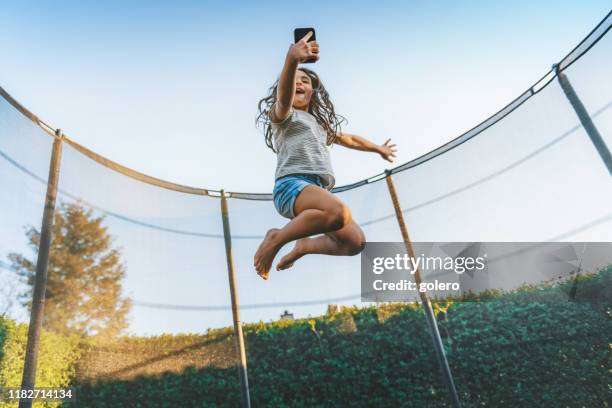 kleines mädchen springt hoch auf trampolin mit handy - trampoline stock-fotos und bilder