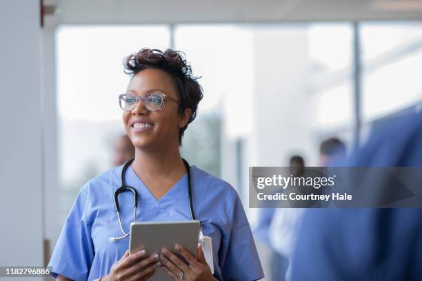 vrouwelijke verpleegkundige of arts glimlacht terwijl staren uit venster in ziekenhuis hal en het houden van digitale tablet met elektronische patiëntendossier - doctor scrubs stockfoto's en -beelden