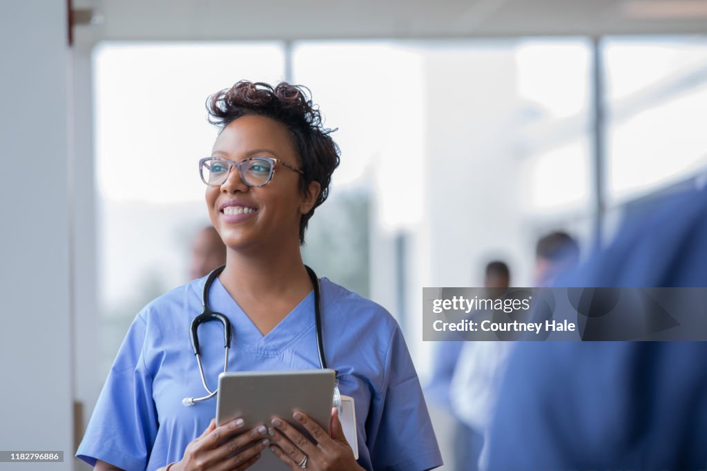Female nurse or doctor smiles while staring out window in hospital hallway and holding digital tablet with electronic patient file