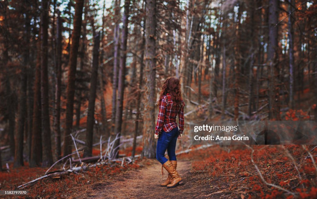 Rear view of young woman in red plaid shirt, jeans, and tan boots standing alone on forest path, autumn