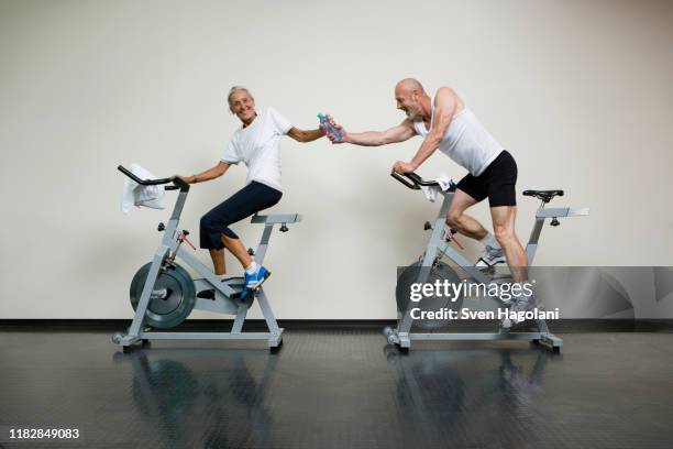 a senior woman and a mature man riding stationary bikes and passing a water bottle - spinning bikes stock pictures, royalty-free photos & images