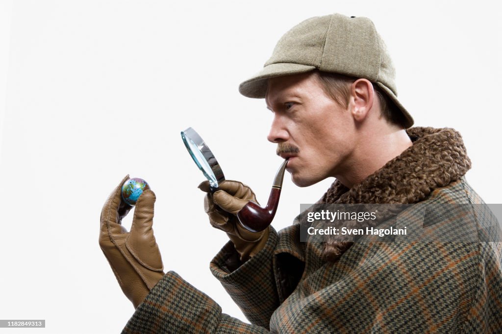 A man dressed up as Sherlock Holmes looking at a tiny globe through a magnifying glass