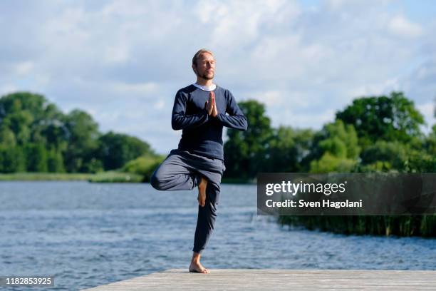 mature man practicing tree pose yoga on pier by lake against sky - tree position stock pictures, royalty-free photos & images