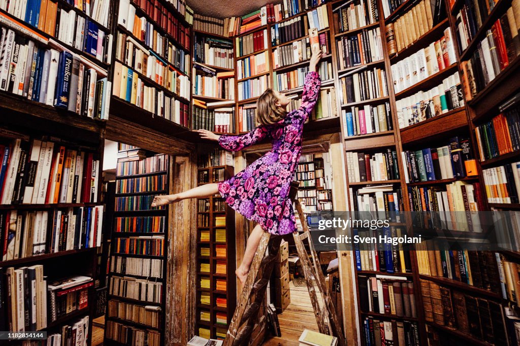 Carefree woman on ladder reaching for book in library