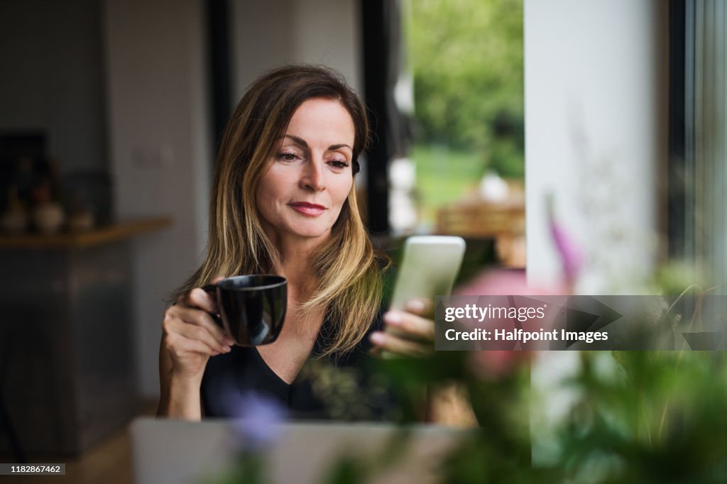 Front view portrait of mature woman with laptop sitting indoors at the table, using smartphone.