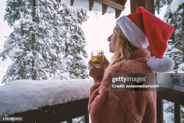 woman with christmas hat on terrace chalet looking at the snow falling with tea cup in hands - terrace british columbia stock pictures, royalty-free photos & images