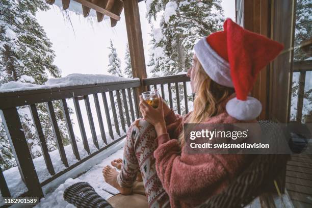 woman with christmas hat on terrace chalet looking at the snow falling with tea cup in hands - terrace british columbia stock pictures, royalty-free photos & images