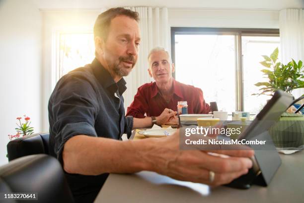 senior gay couple having a lively conversation at breakfast about news on tablet - gay couple stock pictures, royalty-free photos & images