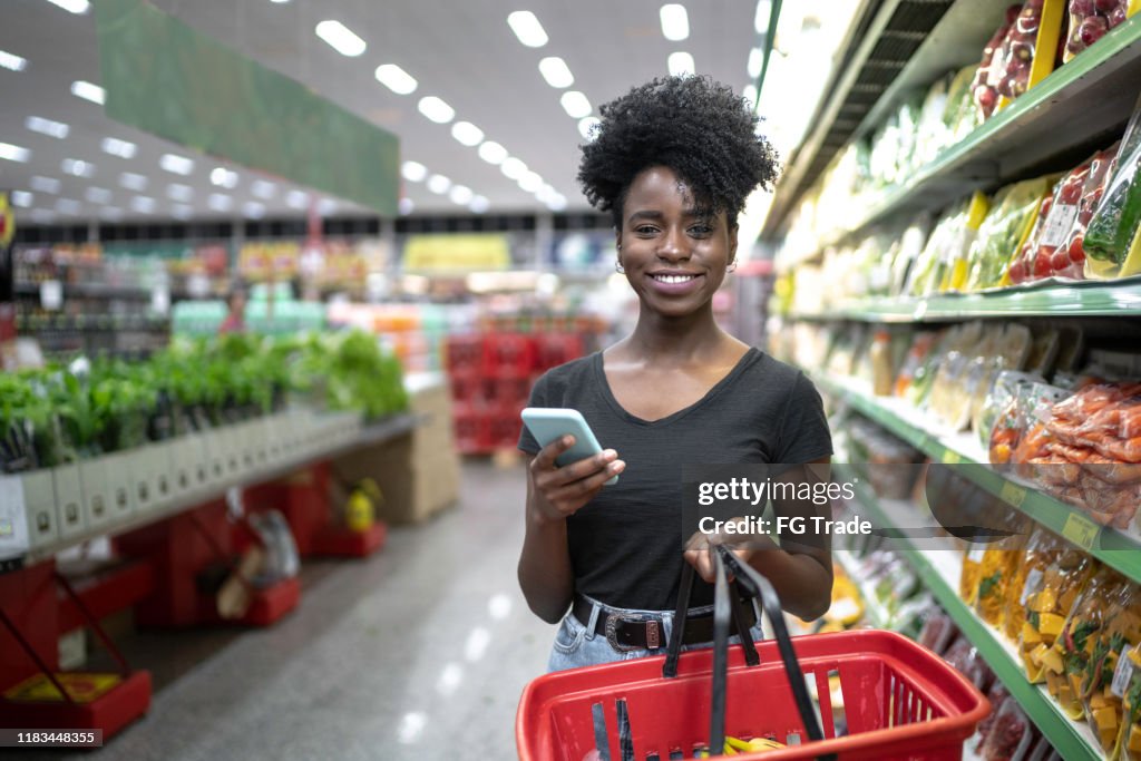 Retrato da mulher de sorriso que usa o telefone e que prende uma cesta de compra no supermercado