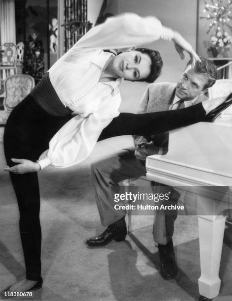 Cyd Charisse , US actress and dancer, holding a ballet pose, standing on one leg with the other leg outstretched and resting on a piano, at which...