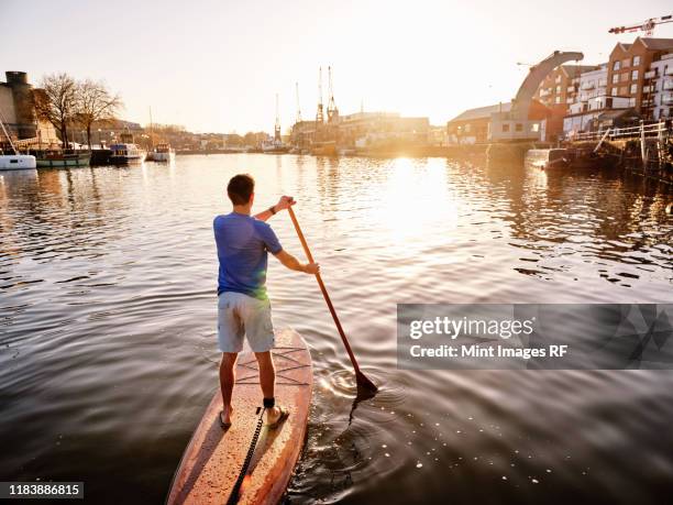 man standing on paddleboard on river at dawn, shot from behind - bristol stock-fotos und bilder